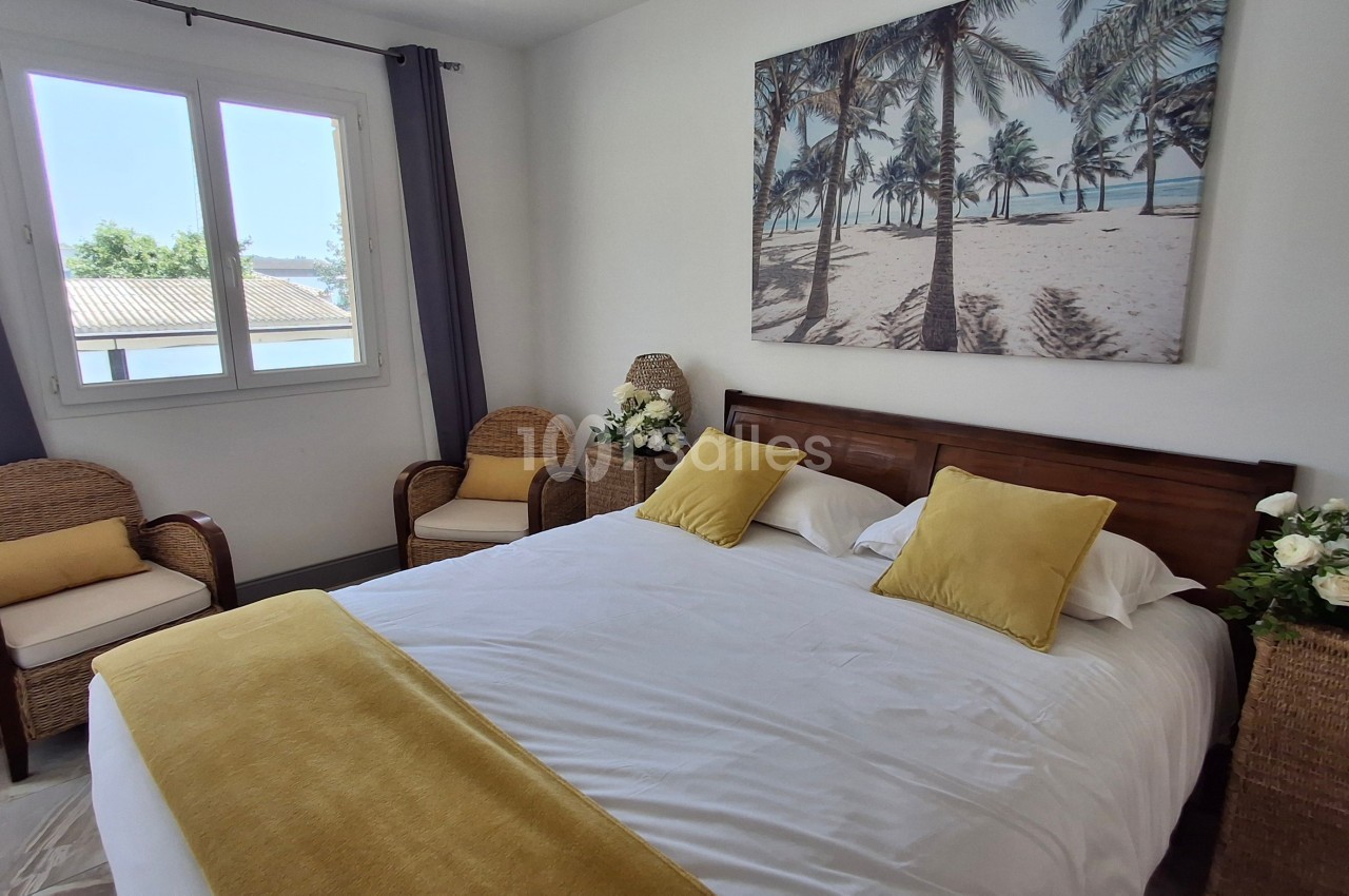Chambre lumineuse avec un lit double, coussins jaunes, fauteuils en rotin et tableau représentant une plage tropicale.