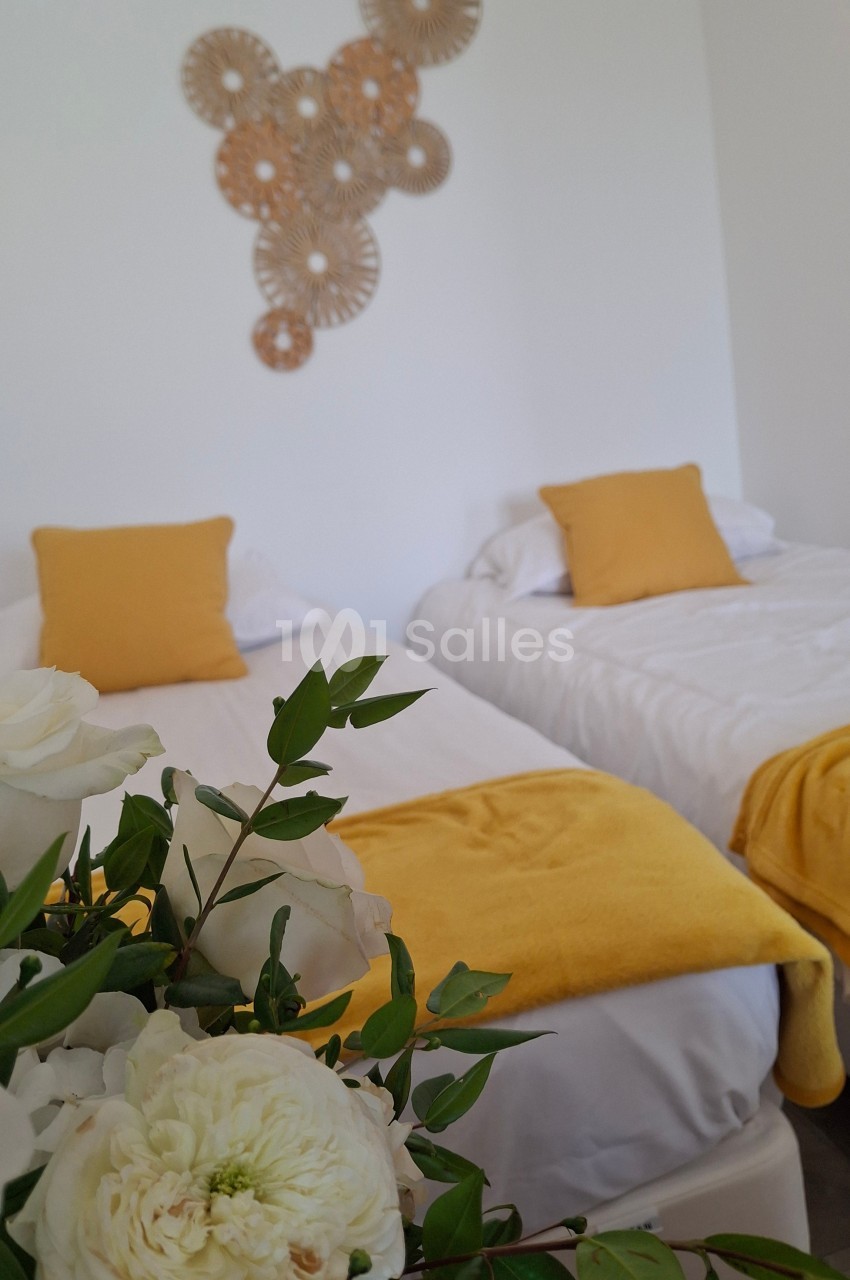 Deux lits simples avec linge blanc et coussins jaunes, décorés de fleurs blanches au premier plan et d'un mur orné.