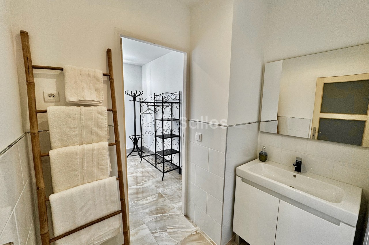 Salle de bain lumineuse avec lavabo, miroir, porte ouverte sur un espace avec porte-manteaux et échelle porte-serviettes.
