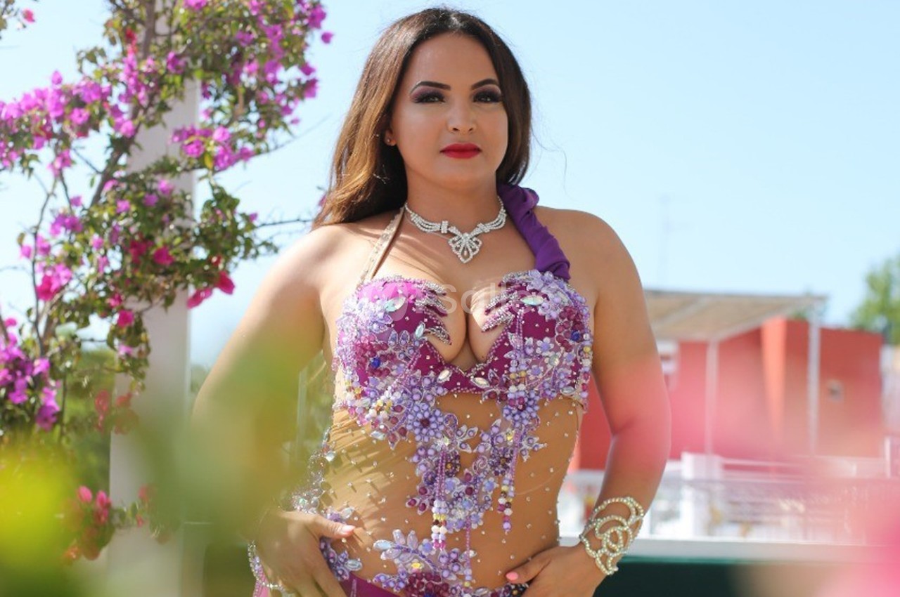 Femme en tenue de danse orientale violette ornée de motifs brillants, posant devant des fleurs et un ciel dégagé.