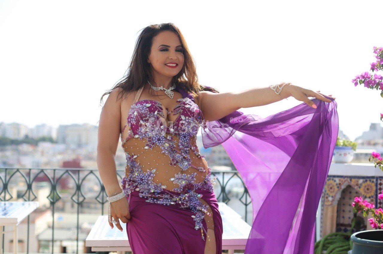 Femme en tenue de danse orientale violette ornée de perles, posant sur une terrasse ensoleillée avec vue urbaine.