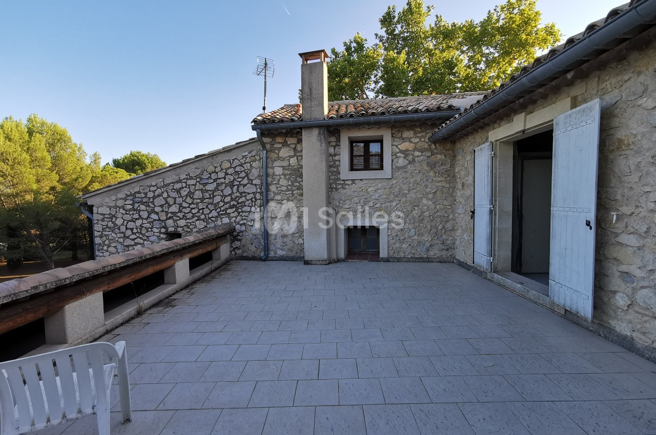 Terrasse en carrelage clair avec vue sur une maison en pierre, entourée de végétation et ciel dégagé.