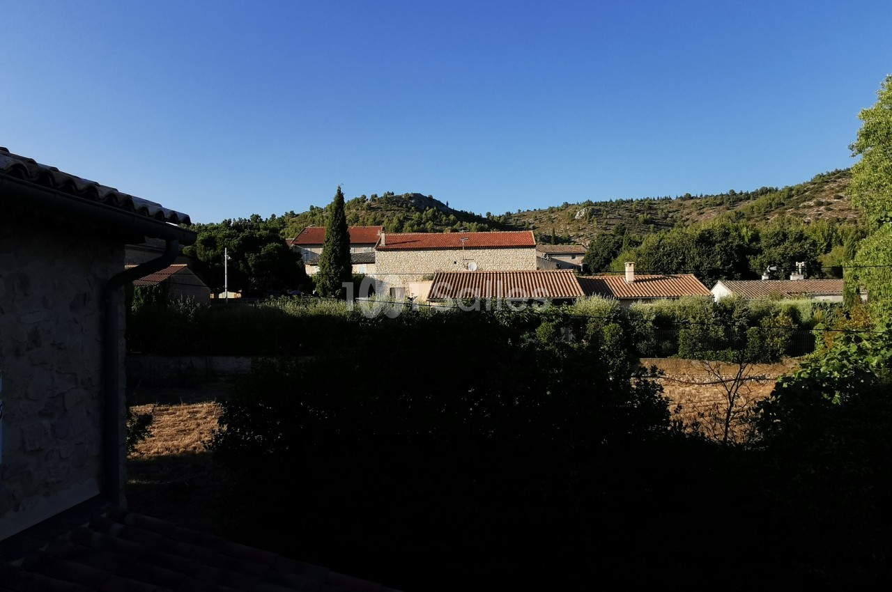 Vue d'un village avec des toits en tuiles rouges, entouré de végétation et de collines sous un ciel bleu clair.