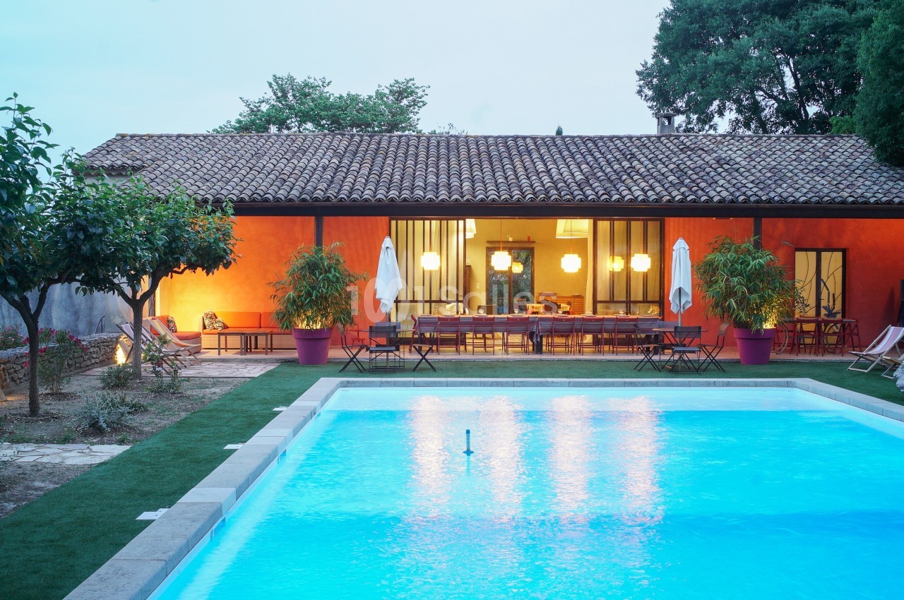 Maison avec façade orange, éclairée en soirée, entourée de végétation, avec une piscine éclairée au premier plan.