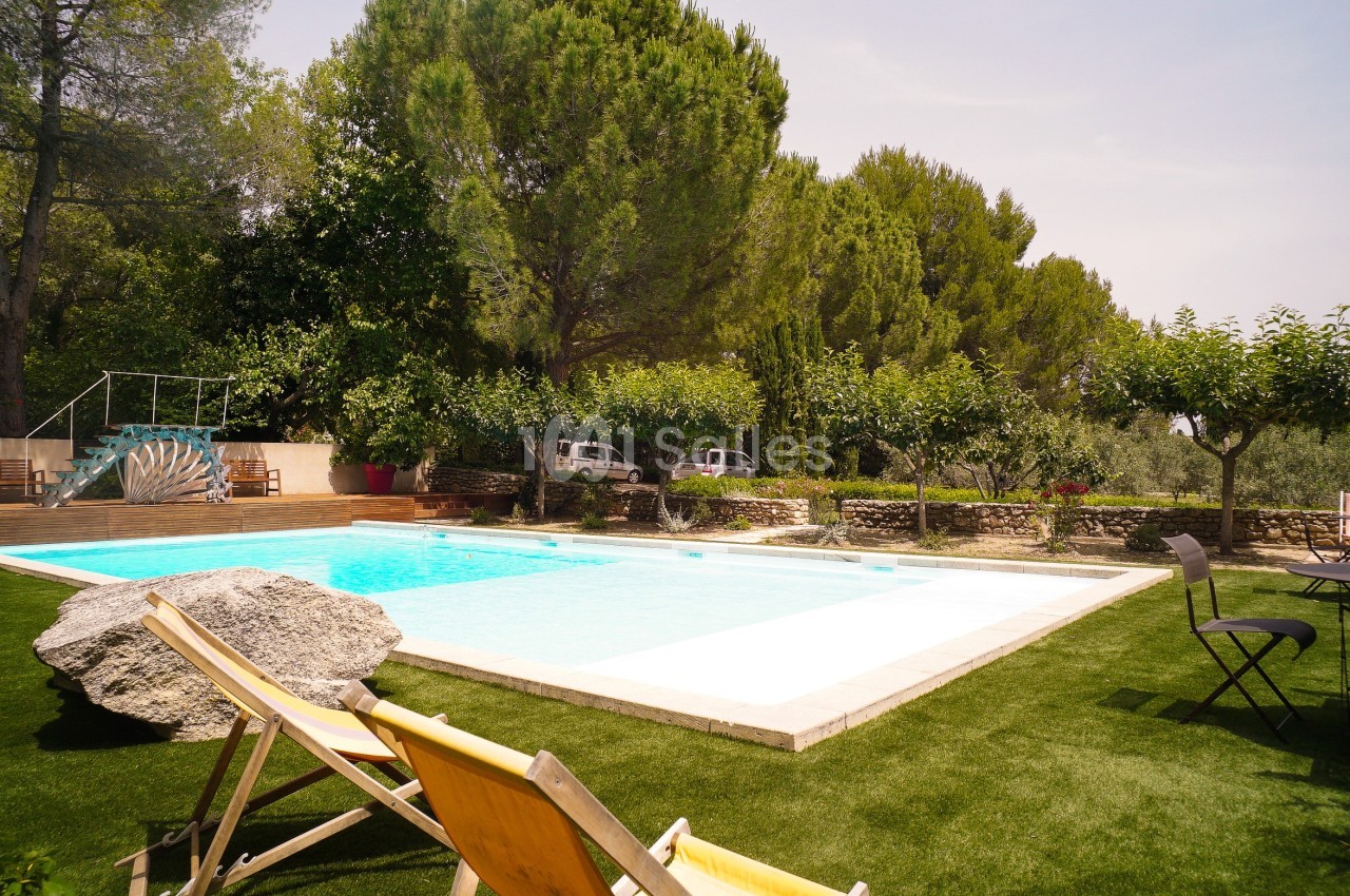 Piscine extérieure entourée de verdure, avec des chaises longues et des arbres sous un ciel dégagé.