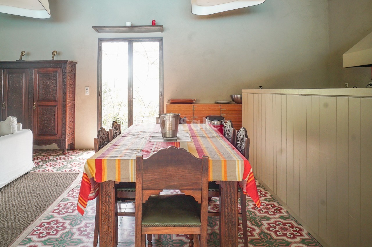 Salle à manger lumineuse avec une grande table recouverte d'une nappe colorée, entourée de chaises en bois.