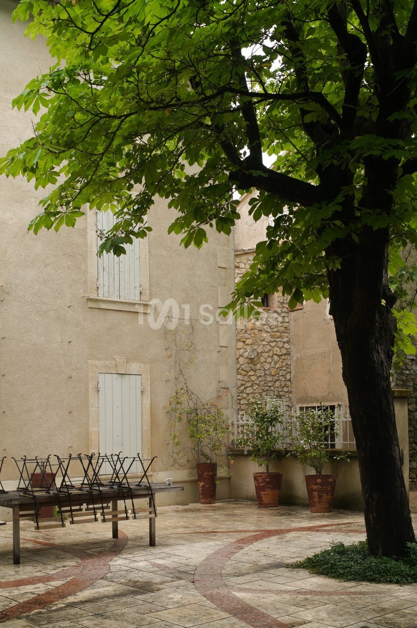 Cour pavée avec un arbre, des chaises empilées, des pots de fleurs et une façade beige aux volets fermés.