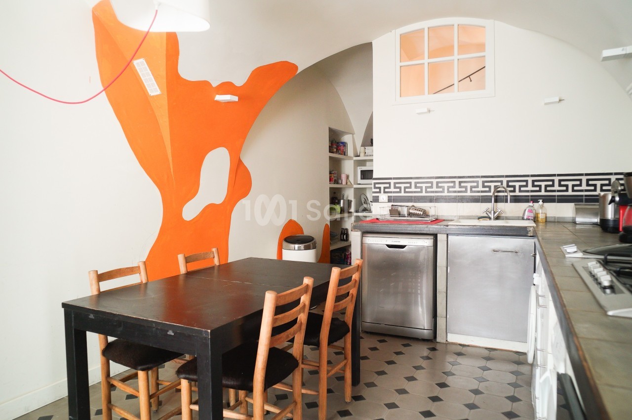 Cuisine avec table en bois, chaises, lave-vaisselle, plan de travail et mur décoratif orange vif.