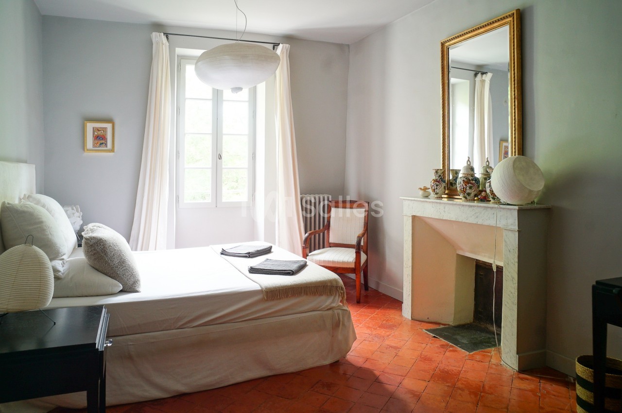Chambre lumineuse avec lit double, cheminée blanche, miroir doré, sol en terre cuite et fenêtre donnant sur l'extérieur.