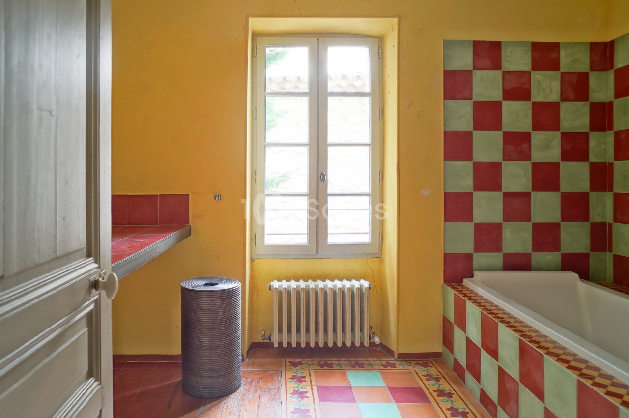 Salle de bain lumineuse avec murs jaunes, baignoire à carreaux rouges et verts, et fenêtre donnant sur l'extérieur.
