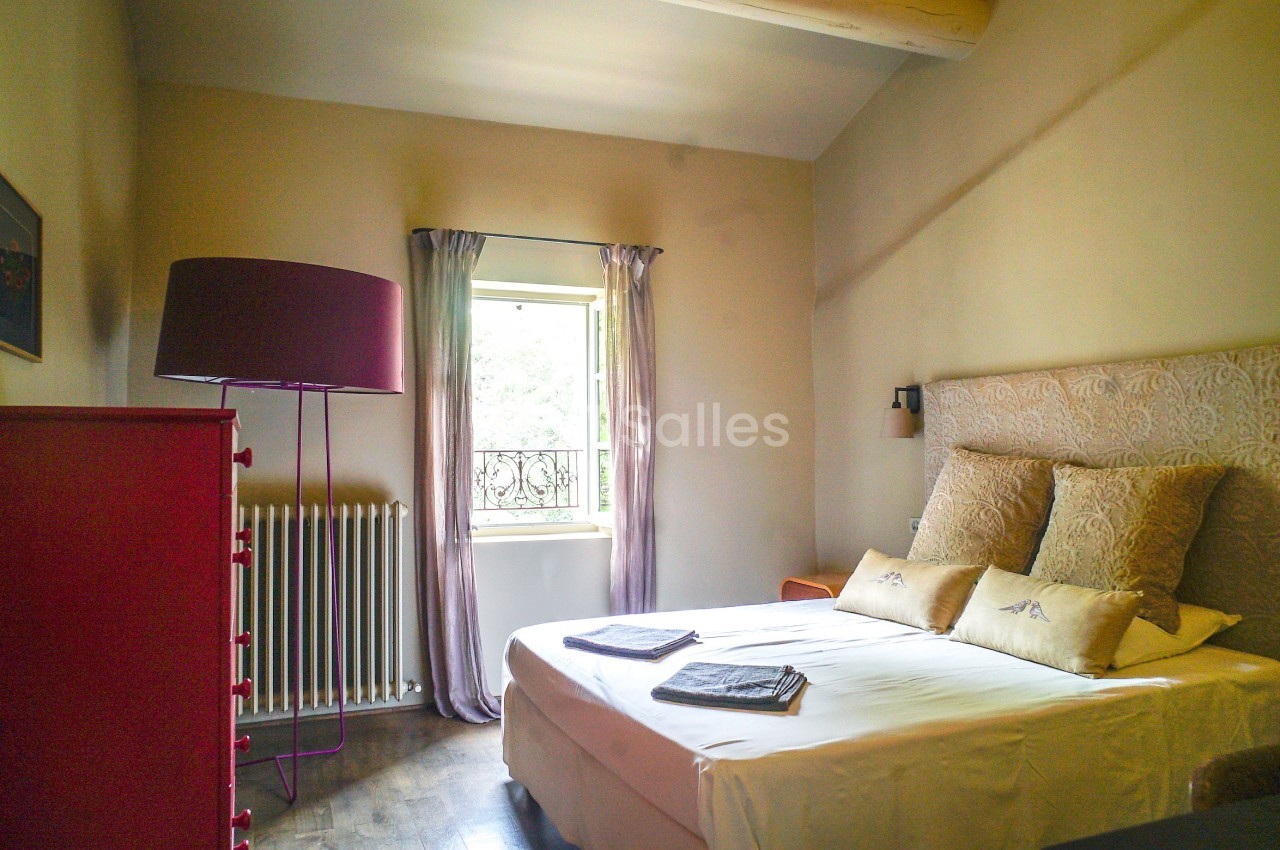 Chambre lumineuse avec lit double, coussins décoratifs, commode rouge, lampe sur pied et fenêtre donnant sur l'extérieur.
