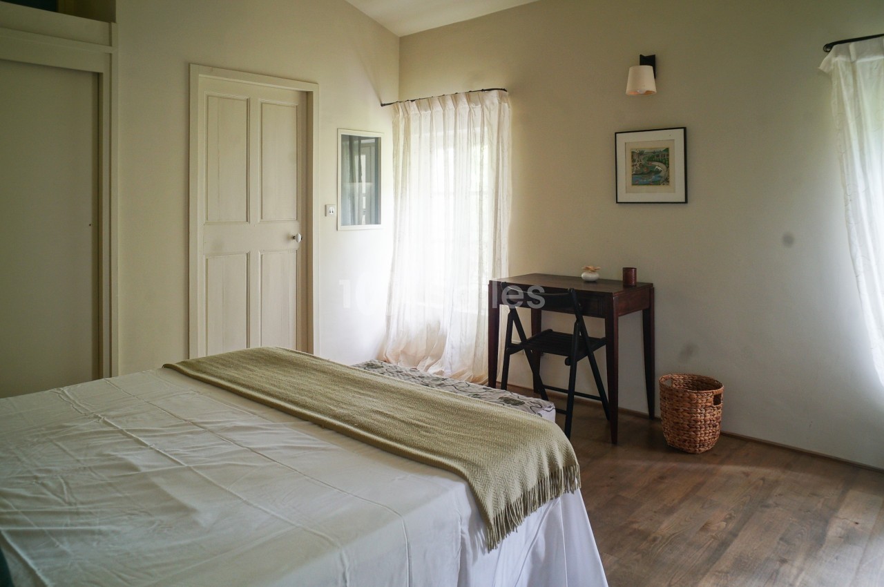 Chambre lumineuse avec lit double, bureau en bois, panier en osier et rideaux blancs laissant passer la lumière naturelle.