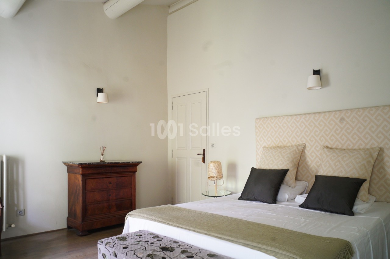 Chambre lumineuse avec lit double, tête de lit beige, commode en bois, chaise et murs blancs épurés.