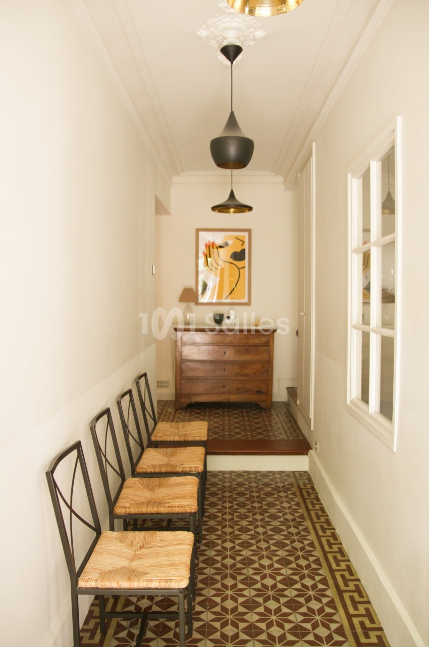 Couloir étroit avec carrelage géométrique, chaises alignées à gauche, commode en bois et tableau au fond.