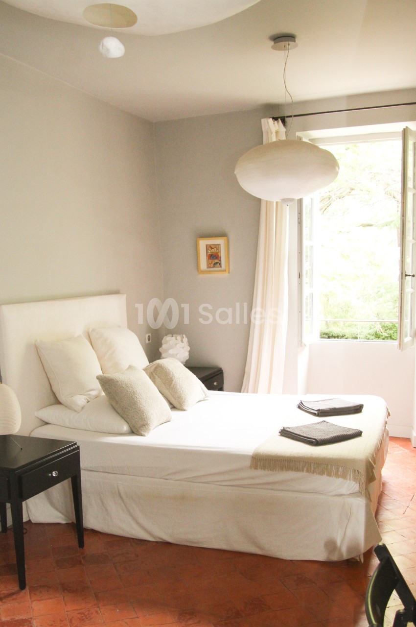 Chambre lumineuse avec lit double, tables de chevet noires, lampe suspendue et fenêtre ouverte sur un jardin.