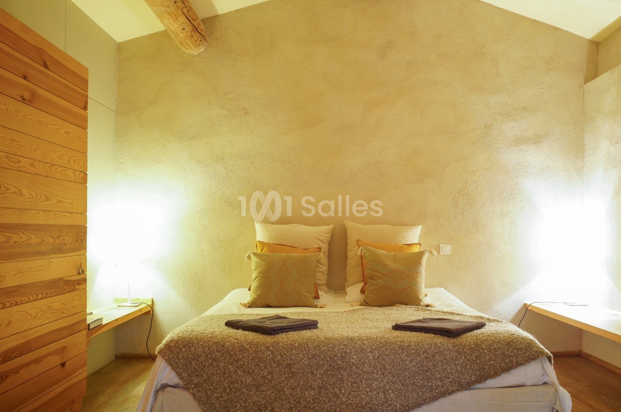 Chambre avec lit double, murs en enduit beige, poutres apparentes et éclairage doux sur des tables de chevet.