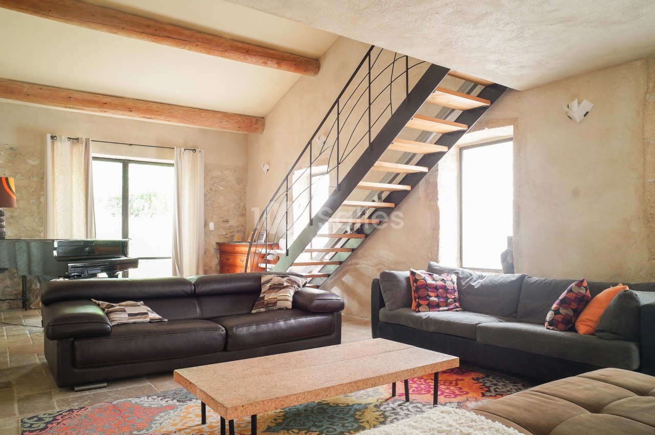Salon lumineux avec murs en pierre, canapé en cuir, tapis coloré, escalier en métal et piano près d'une fenêtre.