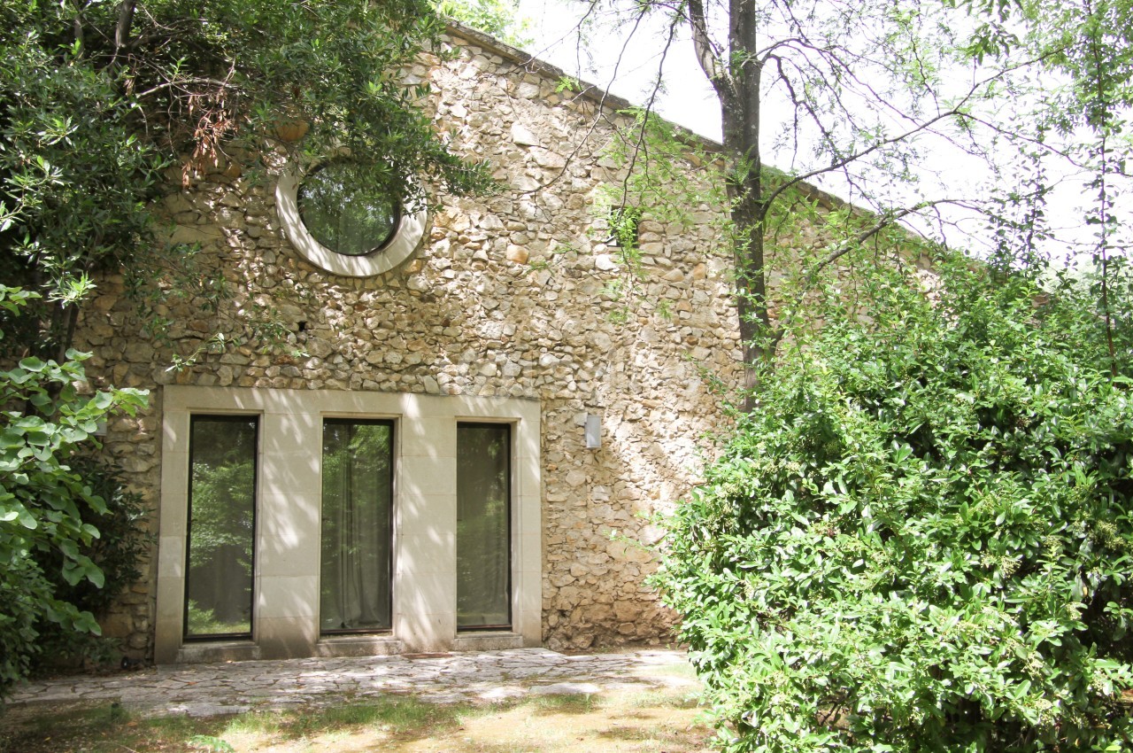 Façade en pierre d'une maison avec une grande porte vitrée et une fenêtre ronde, entourée de végétation.