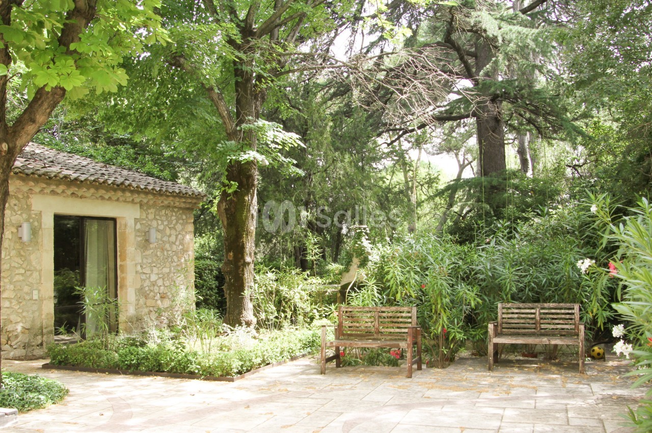 Jardin arboré avec une petite maison en pierre, deux bancs en bois et un sol en dalles claires.