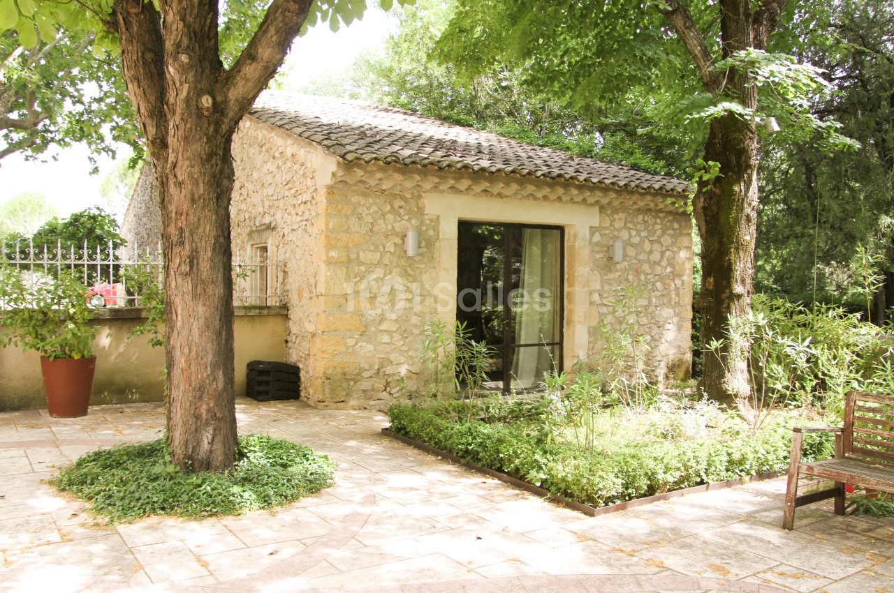 Petite maison en pierre entourée de verdure, avec une terrasse pavée ombragée par des arbres.