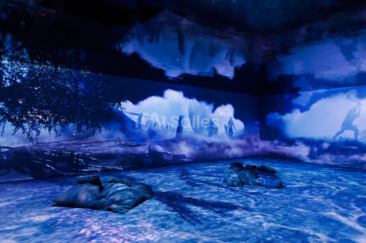 Projection immersive avec des paysages bleutés, des silhouettes humaines et des sacs posés au sol dans une salle sombre.
