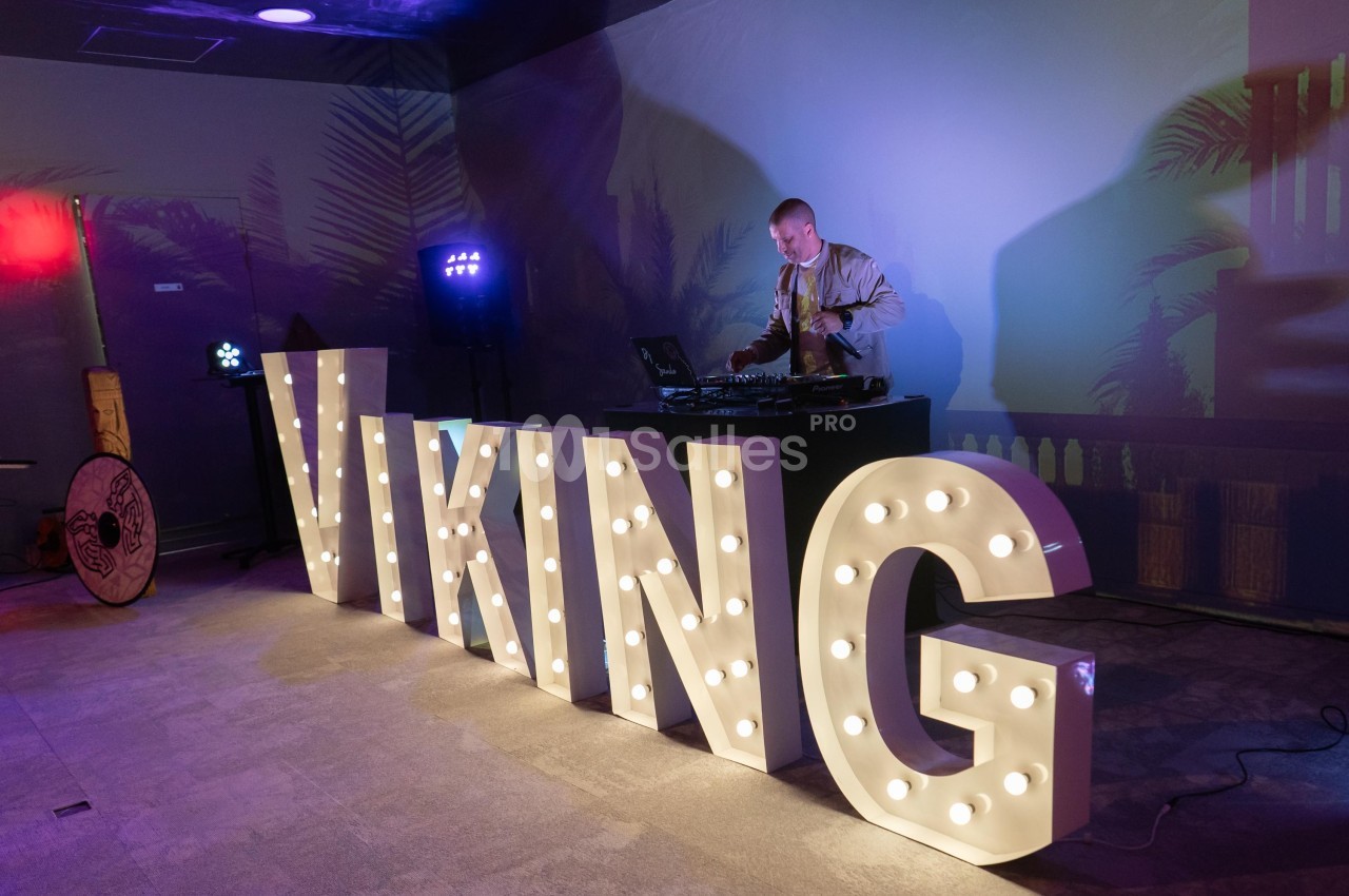 Un DJ mixe de la musique derrière des lettres lumineuses formant le mot ’VIKING’ dans une ambiance tamisée.