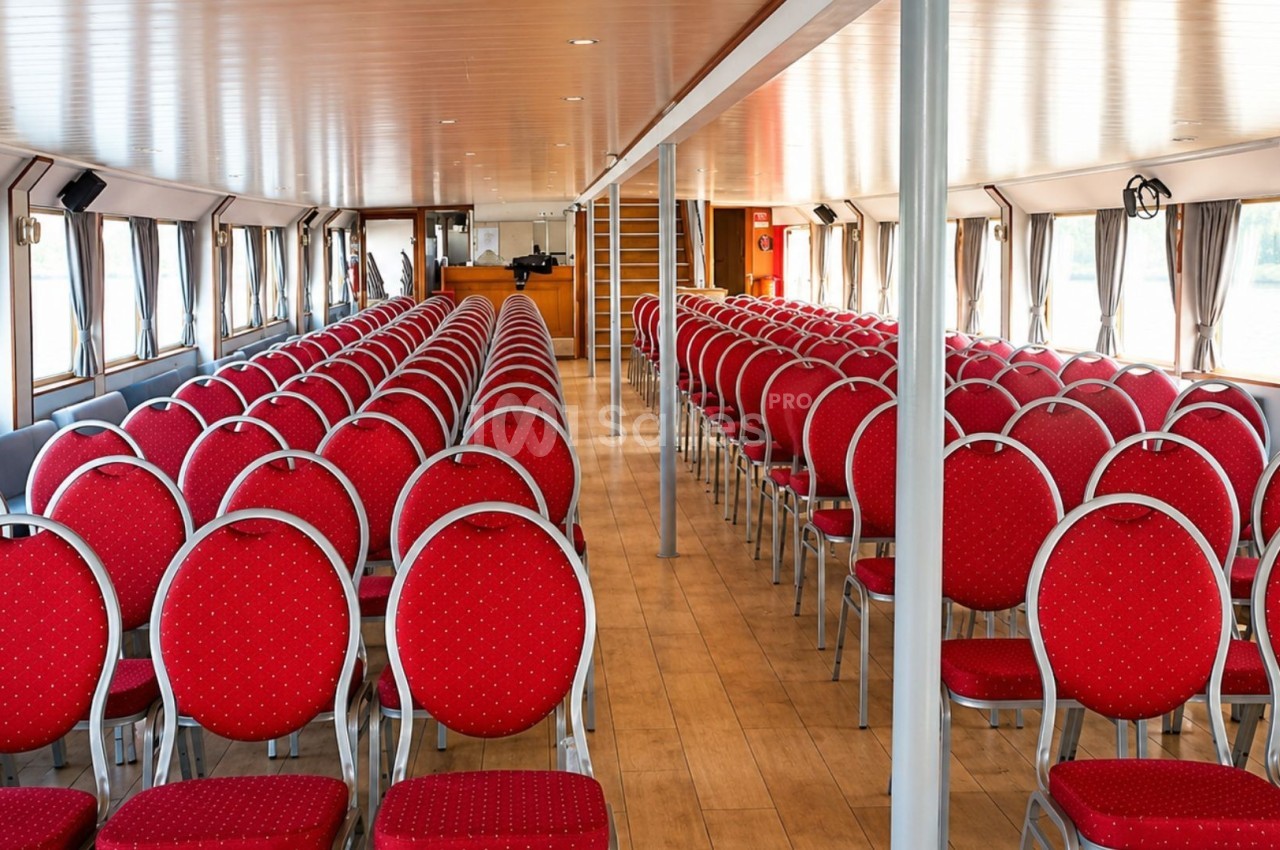 Salle avec rangées de chaises rouges alignées, éclairée par des fenêtres latérales et un plafond en bois clair.