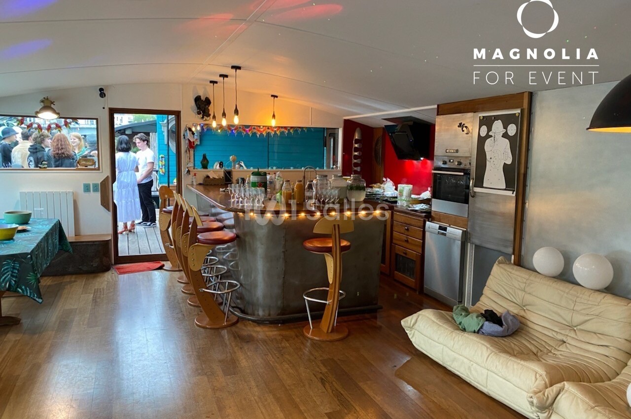 Intérieur d'une salle avec bar, tabourets, canapé, cuisine équipée et vue sur une terrasse avec des invités.