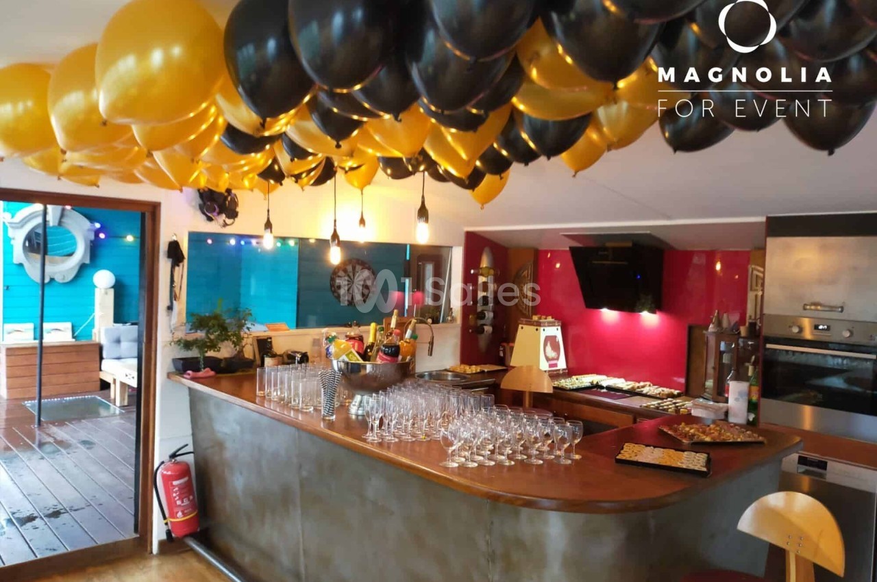 Bar décoré avec des ballons noirs et dorés, verres alignés sur le comptoir, espace festif avec terrasse en arrière-plan.