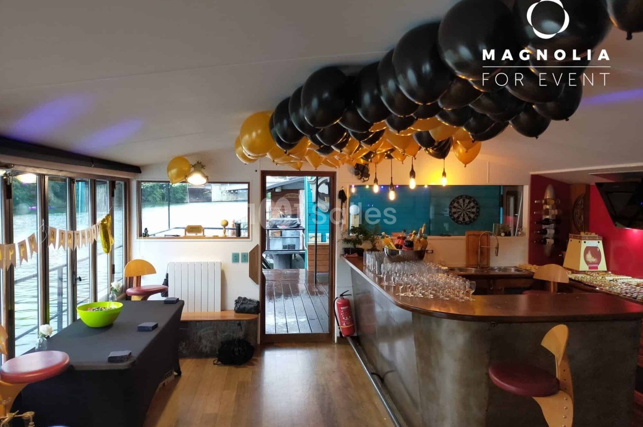 Salle décorée pour un événement avec ballons noirs et dorés, bar équipé et vue sur une terrasse extérieure.