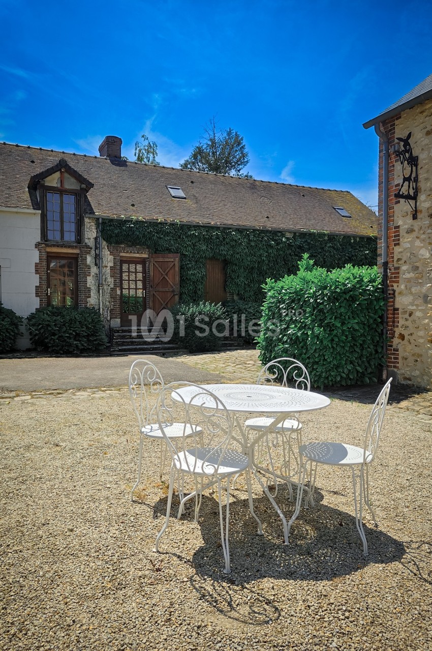Table et chaises blanches en fer forgé sur une cour en gravier, devant une maison en pierre avec façade végétalisée.