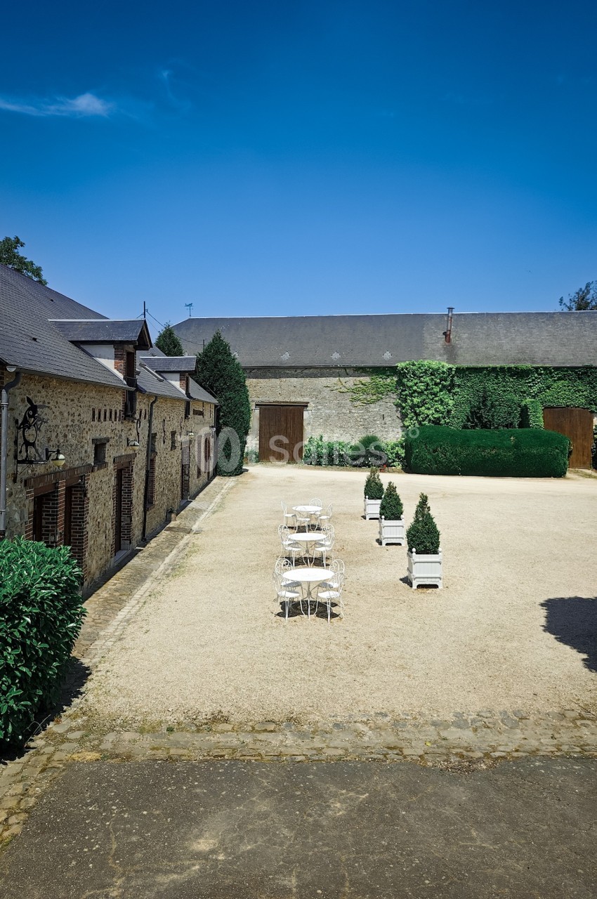 Cour intérieure pavée avec tables et chaises blanches, entourée de bâtiments en pierre et de verdure sous un ciel bleu.