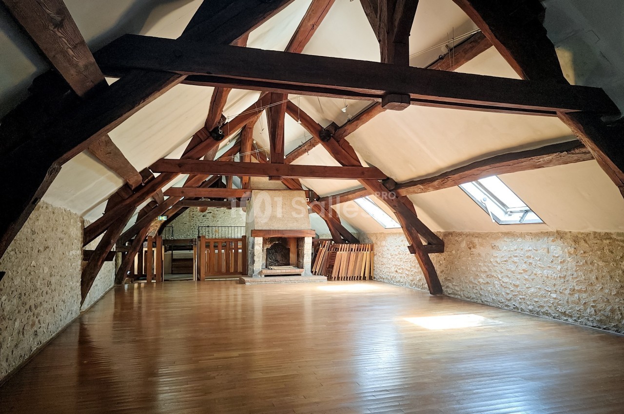 Grande pièce sous combles avec poutres apparentes, parquet en bois, murs en pierre et cheminée centrale.