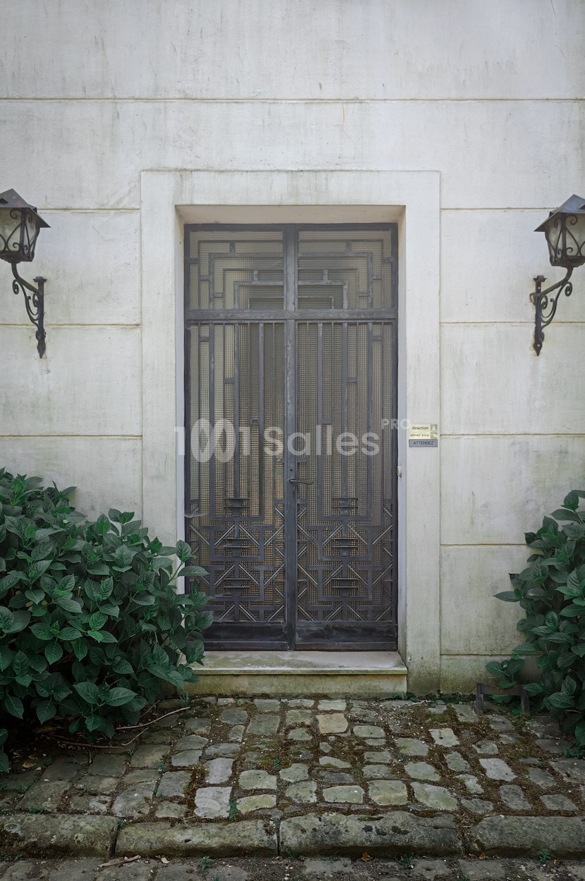 Porte en fer forgé au design géométrique, encadrée par deux lampes murales et des arbustes sur un sol pavé.