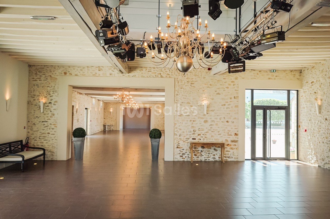 Salle lumineuse avec murs en pierre, lustres suspendus et éclairages, ouverte sur l'extérieur par une porte vitrée.