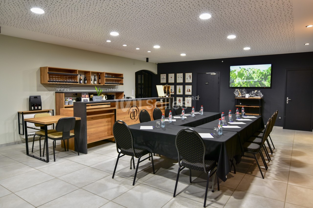 Salle de réunion avec une grande table rectangulaire, chaises noires, écran mural et coin bar en arrière-plan.