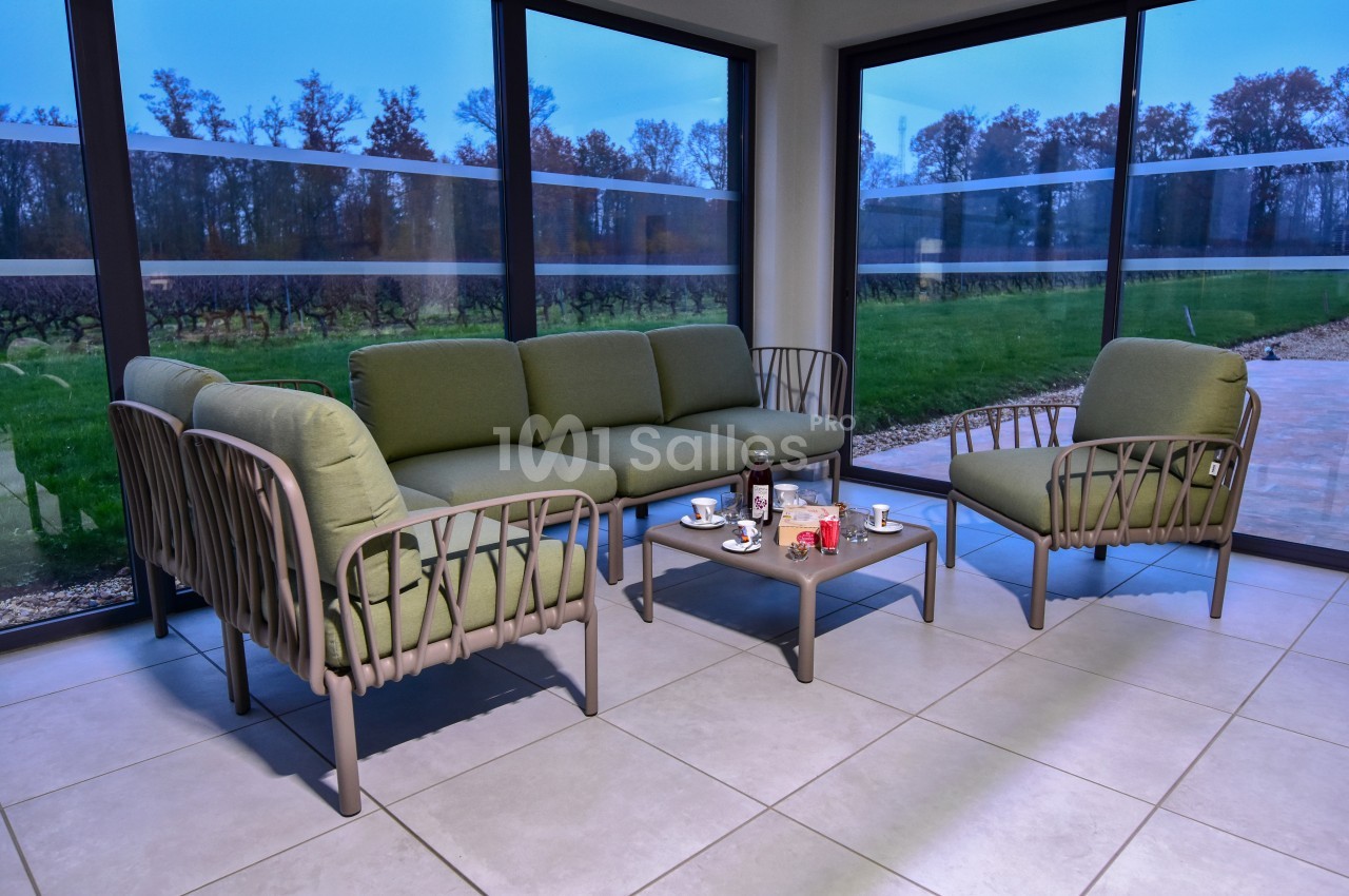 Salon de jardin avec fauteuils et canapé verts autour d'une table basse, situé dans une véranda avec vue sur un jardin.