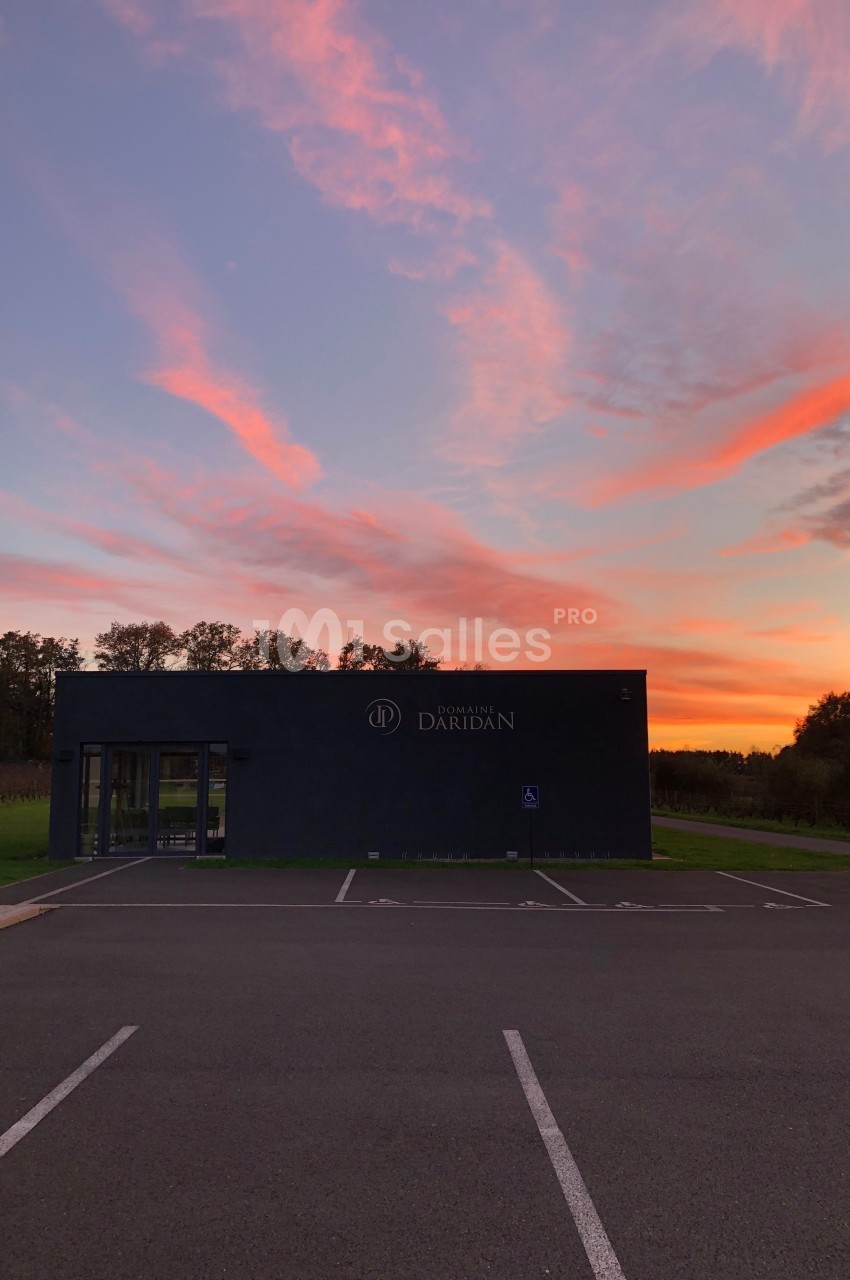 Bâtiment moderne sombre devant un parking vide, au coucher du soleil avec un ciel aux teintes rosées et orangées.