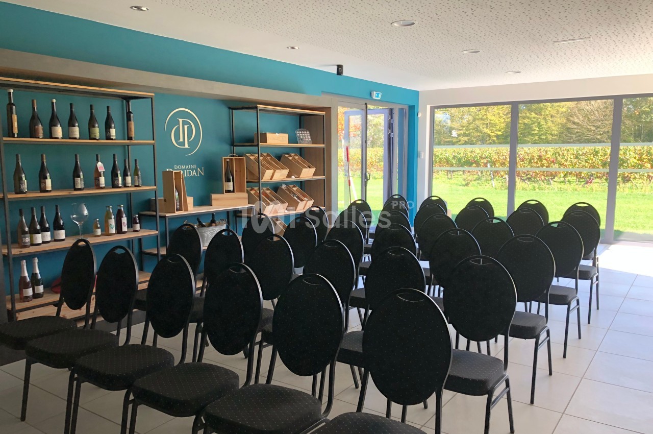 Salle lumineuse avec des chaises alignées face à une étagère contenant des bouteilles et des livres, vue sur un jardin.
