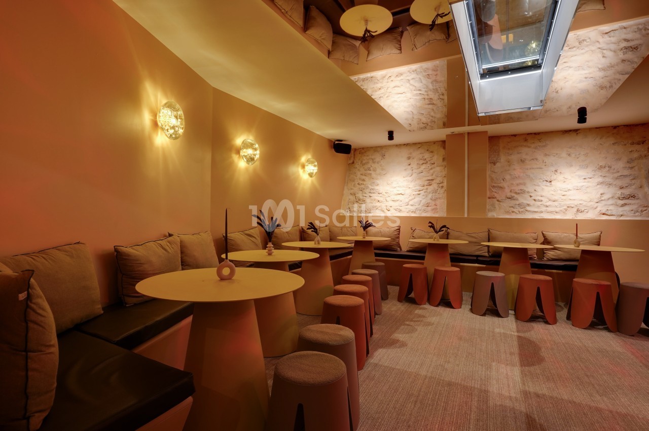 Salle lumineuse avec murs en pierre, banquettes beige, tables rondes et tabourets dans une ambiance chaleureuse.