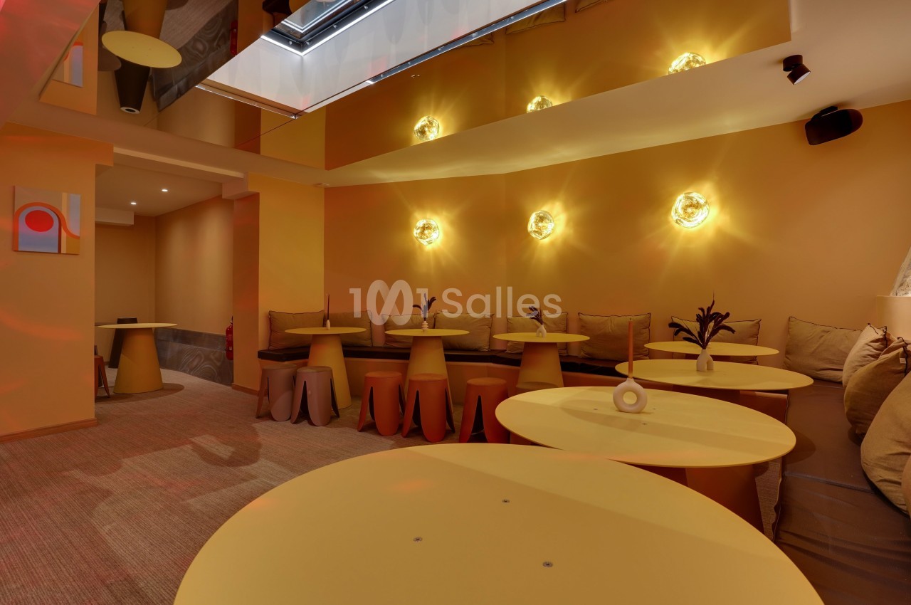 Salle lumineuse avec murs jaunes, tables rondes assorties et banquettes garnies de coussins sous des appliques murales.