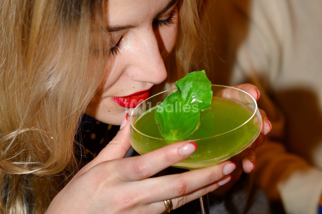 Une femme aux cheveux blonds sent un cocktail vert garni d'une feuille, tenant délicatement le verre.