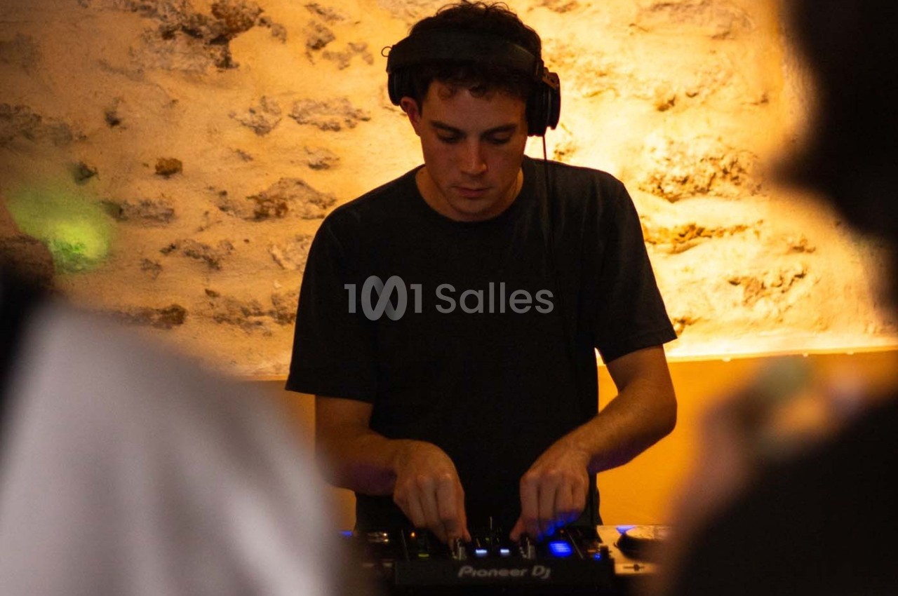 Un DJ mixe de la musique sur une console dans un environnement éclairé de tons chauds avec un mur en pierre en arrière-plan.