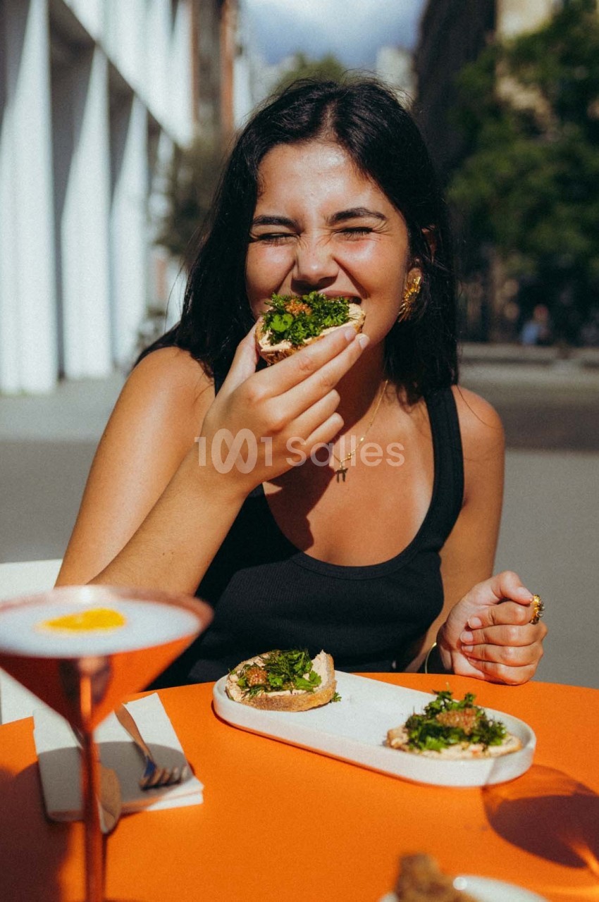 Femme souriante mangeant une tartine garnie de légumes verts à une table en extérieur par une journée ensoleillée.