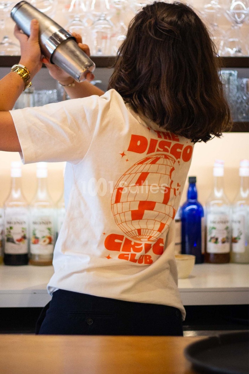 Une personne de dos prépare un cocktail avec un shaker dans un bar, portant un t-shirt blanc à motif orange.