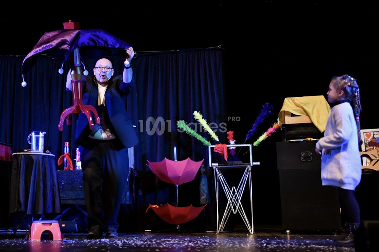 Un magicien sur scène réalise un tour avec un parapluie coloré devant une jeune spectatrice attentive.