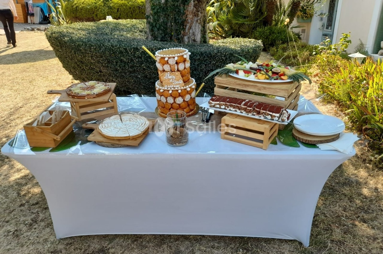Table de buffet en extérieur avec pâtisseries, fruits frais, croquembouche et plateaux en bois sur une nappe blanche.