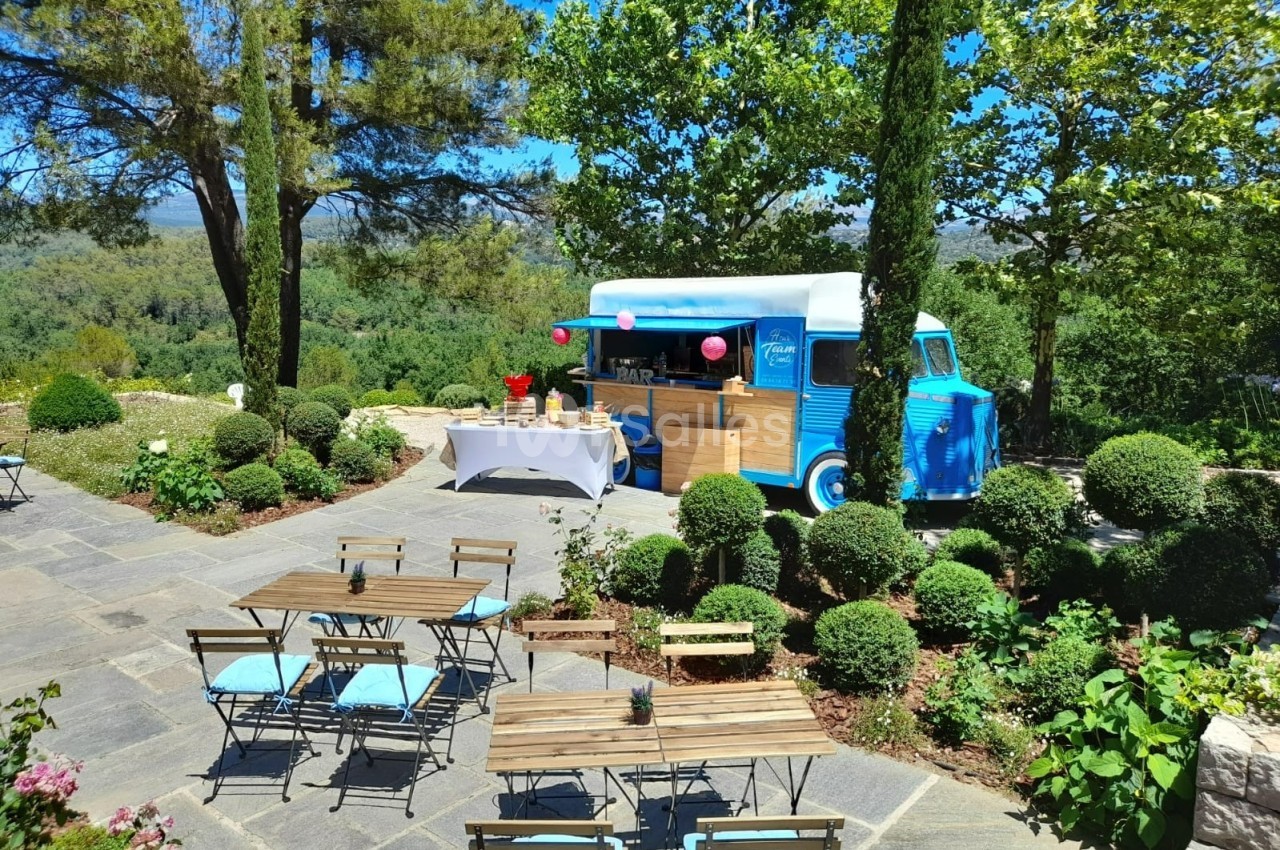 Camion bleu aménagé en bar, stationné dans un jardin avec tables en bois et chaises, entouré de verdure.