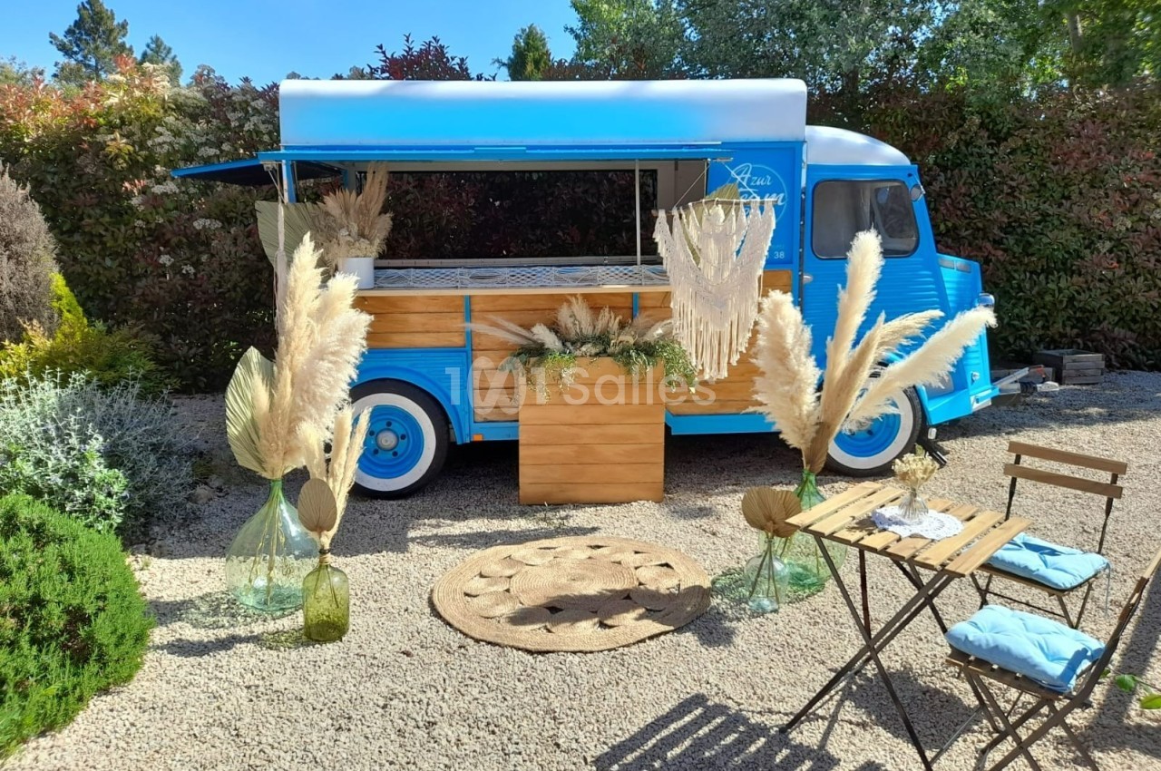 Camion bleu aménagé en food truck, entouré de décorations en pampas, avec table et chaises en bois sur un sol gravillonné.