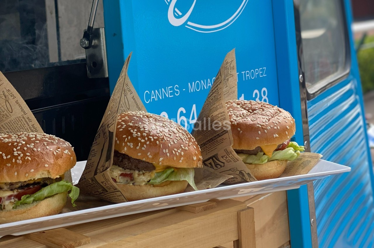 Trois burgers garnis de salade, tomate et fromage, servis sur un plateau devant un food truck bleu.
